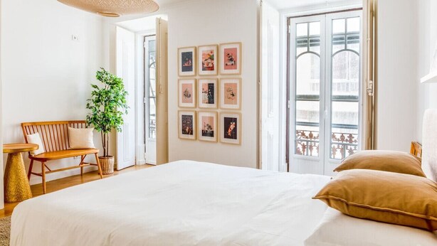 A bedroom with a large bed, a wooden bench, a potted plant, and a wall with framed pictures.