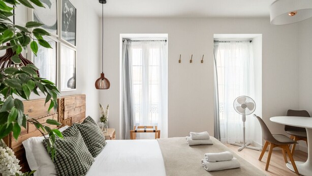 A modern bedroom with a wooden headboard, a large bed with green and white bedding, two windows with sheer curtains, a bedside table with a lamp, and a small desk with a chair. - Elegant Luxury Old Town Studio-apt by Lisbeyond (Lisboa)
