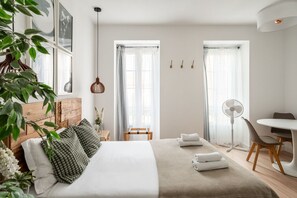 Une chambre moderne avec une tête de lit en bois, un grand lit avec une literie verte et blanche, deux fenêtres avec des rideaux légers, une table de chevet avec une lampe et un petit bureau avec une chaise.