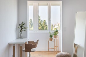 A bright room with a large window, a white desk, a wooden chair, and two wicker side tables with plants. - Spacious Apt Campo D`Ourique by Lisbeyond (Lisboa)