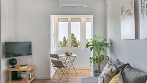 Ett modernt vardagsrum med en platt-TV, ett litet träbord med stolar, en krukväxt och en grå soffa.