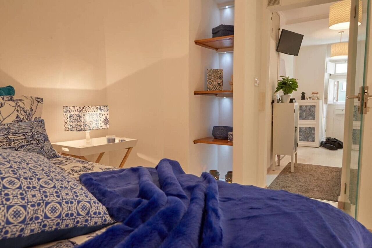 Une chambre à coucher avec un lit, une table de chevet, une lampe et une couverture bleue. On y trouve des étagères ornées d’objets décoratifs et un téléviseur fixé au mur.
