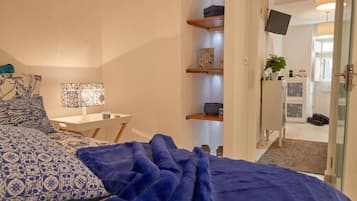 Une chambre à coucher avec un lit, une table de chevet, une lampe et une couverture bleue. On y trouve des étagères ornées d’objets décoratifs et un téléviseur fixé au mur.