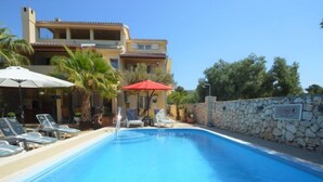 A swimming pool with lounge chairs and umbrellas, surrounded by a stone wall and a building with multiple balconies.