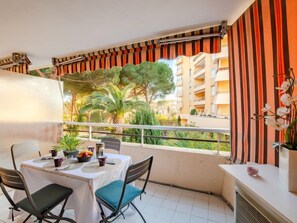 Varanda com mesa posta para dois, cadeiras, vaso de planta e vista para um prédio residencial e árvores.