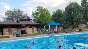 A swimming pool with multiple people in the water, a covered area, and a building in the background. - Spacious mobile home with large kitchen/living area and porch with barbeque. (Longmont)