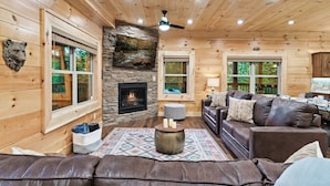 A cozy cabin interior with a stone fireplace, wooden walls, and a leather sofa. - Luxury Gatlinburg Cabin w/ Indoor Pool + Theater + 2 Hot Tubs | Sleeps 24 (Gatlinburg)