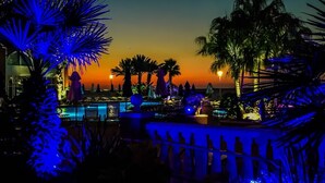 Un espace piscine agrémenté de palmiers et de parasols, un coucher de soleil en arrière-plan et une clôture ornée de silhouettes.