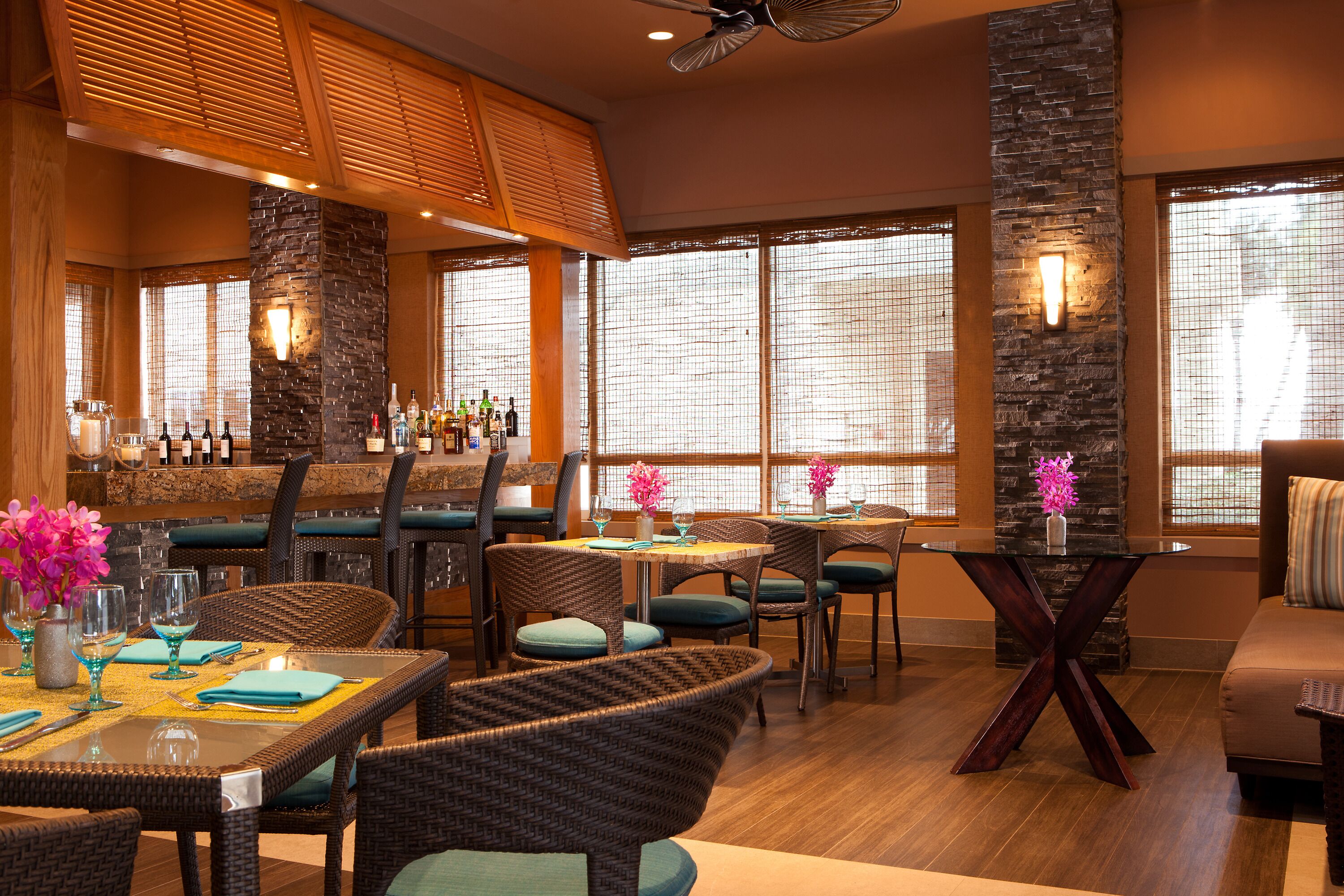 A restaurant with wicker furniture, a bar area, and large windows with blinds.