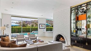 A modern living room with a fireplace, a pool view, and a glass cabinet with decorative items. - Mod Blue Agave - Stunning S Palm Springs Mid-Centu (Palm Springs)