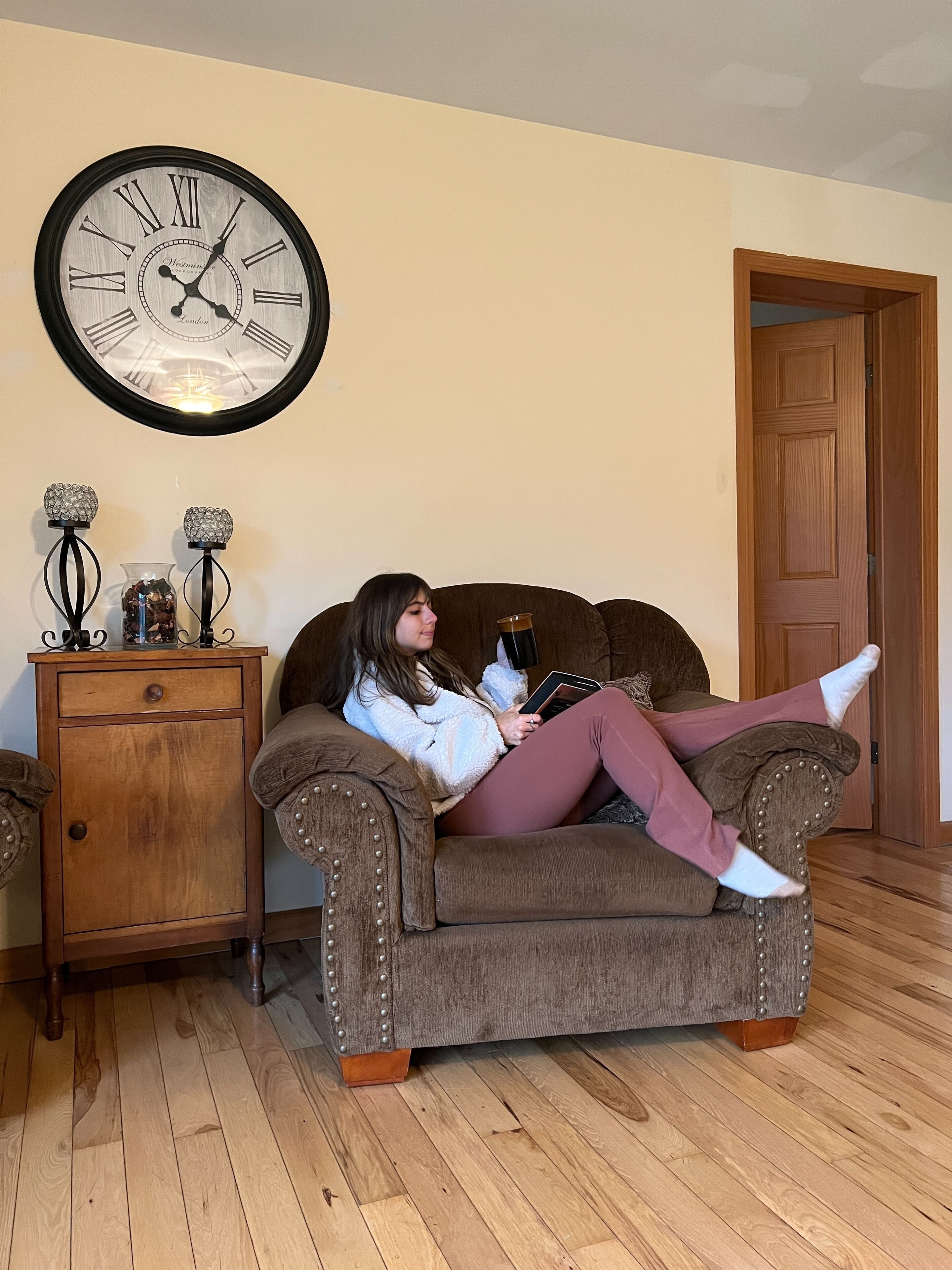 A person sitting on a brown upholstered armchair with a wooden base, reading a book. A large wall clock with Roman numerals is mounted above the armchair.