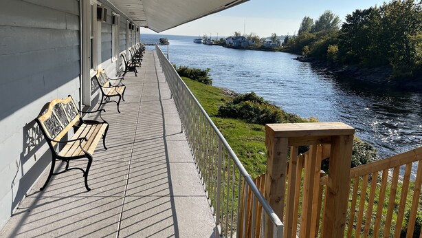A covered walkway with benches overlooking a river, with a view of the river and greenery. - Old Mill Motel (Blind River)
