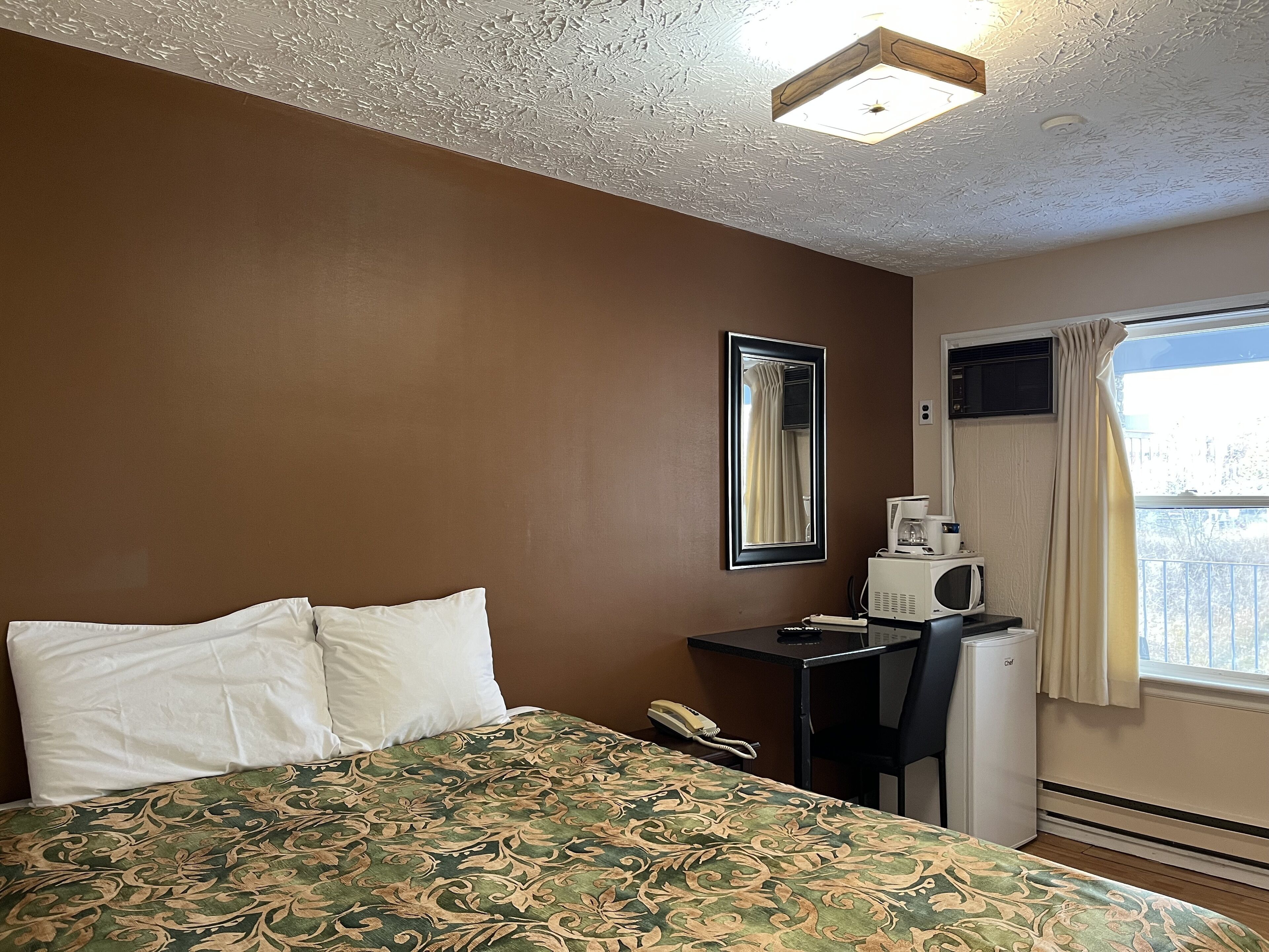A hotel room with a bed, a desk with a telephone, a coffee maker, and a window with curtains.