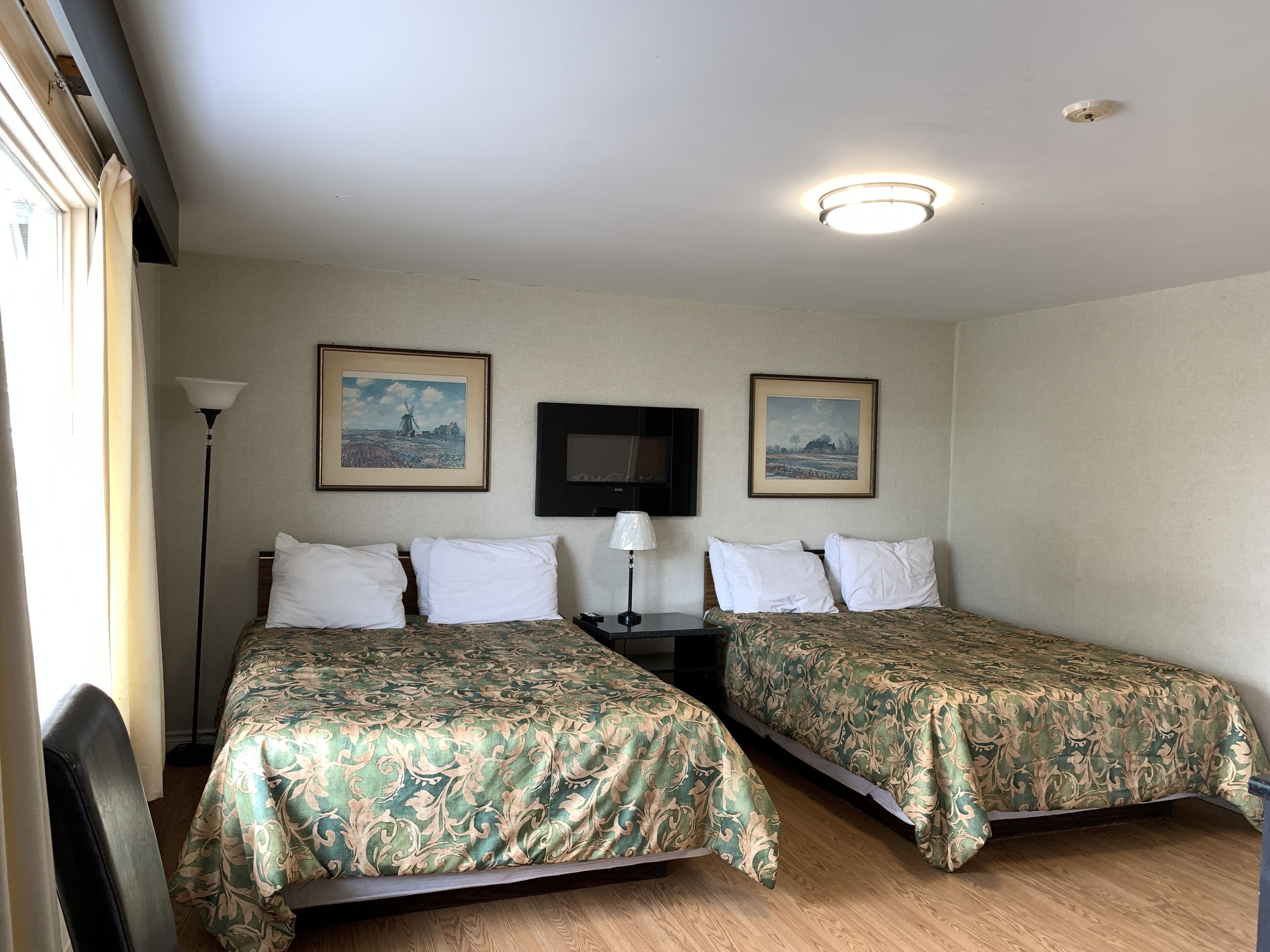 A hotel room with two single beds, a flat-screen TV, a standing lamp, and two framed pictures on the wall.