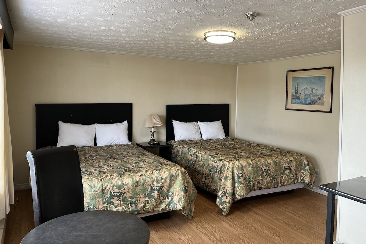 A hotel room with two beds, a small table, a lamp, and a painting on the wall.