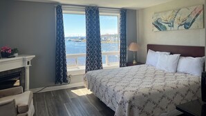 Une chambre d’hôtel avec un lit, une table de chevet avec une lampe, une cheminée, une vue sur l’eau et un tableau au mur.