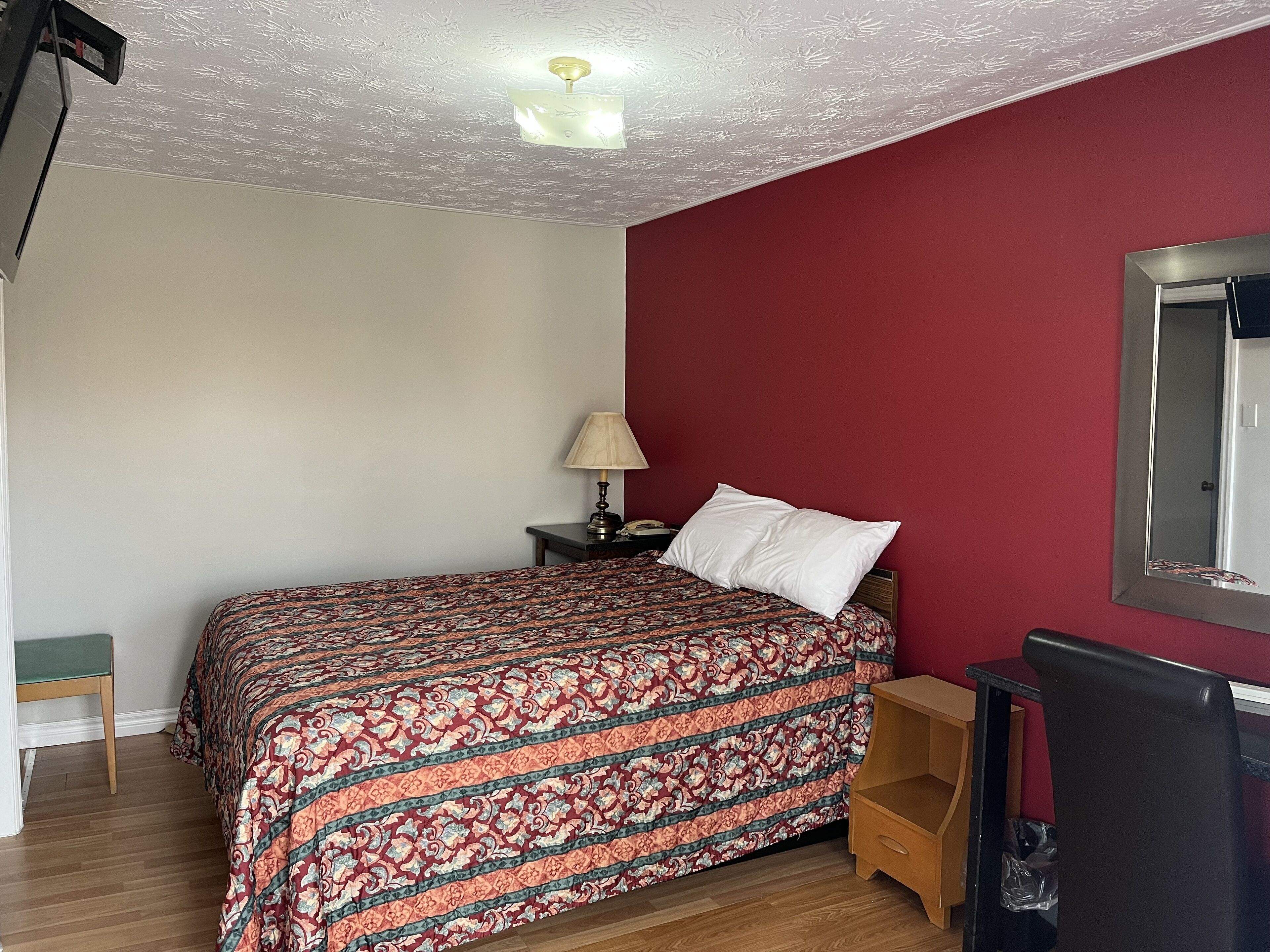 A bedroom with a patterned bedspread, a nightstand with a lamp, a wooden chair, and a mirror on the wall.