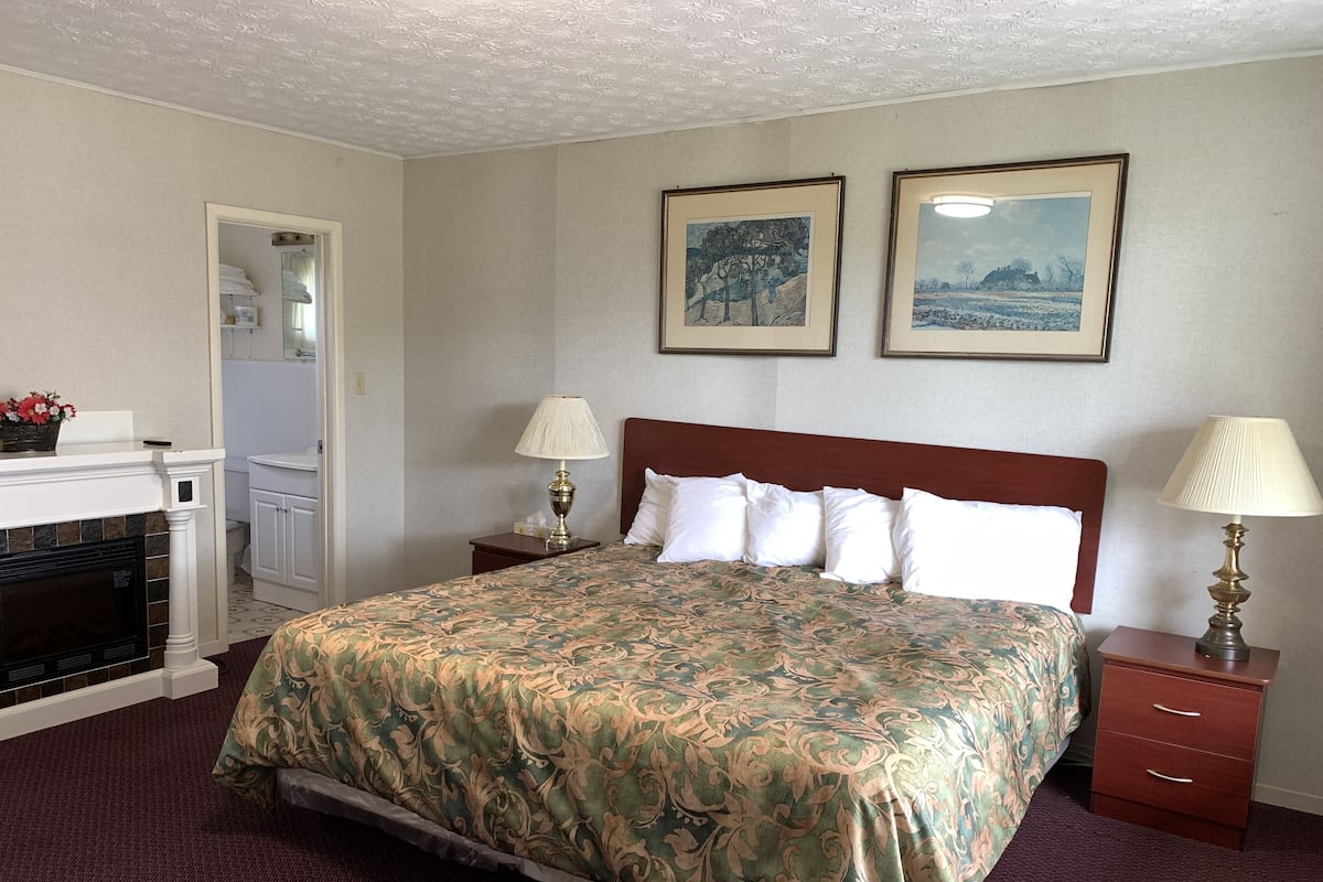 A hotel room with a bed, two bedside lamps, a nightstand, a fireplace, and two framed pictures on the wall.