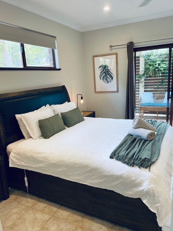 Una cama bien hecha con almohadas y una manta, una lámpara de noche y una planta enmarcada en la pared.
