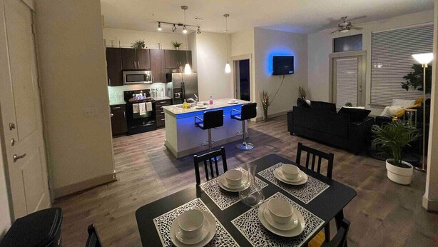 Cocina moderna con isla central, comedor con una mesa dispuesta para cuatro personas y sala de estar con sofá y televisor.