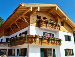 A two-story building with wooden balconies adorned with flowers, green shutters, and a sloped roof. - Steinröschen Vacation Apartment, 30 m², 1 separate bedroom   (Reit im Winkl)
