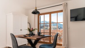 A dining area with a wooden table, two chairs, a potted plant, and a view of mountains through a large window. - Apartment Enzian 32 sqm with balcony, 1 separate bedroom (Reit im Winkl)