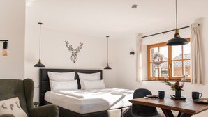 A modern bedroom with a bed, a dining table, and a chair. There is a window with curtains and a view of buildings outside. - Apartment Enzian 32 sqm with balcony, 1 separate bedroom (Reit im Winkl)