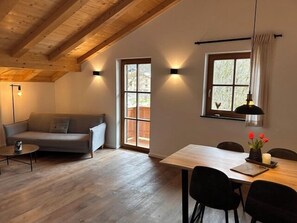 A cozy room with a wooden ceiling, a sofa, a dining table with chairs, and two windows with curtains. - Vacation apartment Alpenmohn 40 sqm, 1 separate bedroom (Reit im Winkl)