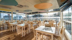 A spacious dining area with multiple tables and chairs, large windows offering views of the cityscape, and a modern ceiling design with circular light fixtures. - Bossa Jurere Hotel (Florianópolis)