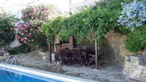 Zona junto a la piscina con un muro de piedra, una mesa y sillas de madera, y un gato.