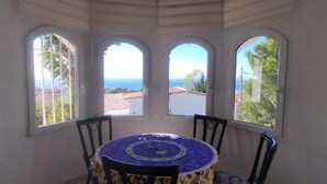 Una habitación con una mesa, sillas, tres ventanas arqueadas y vista al mar.