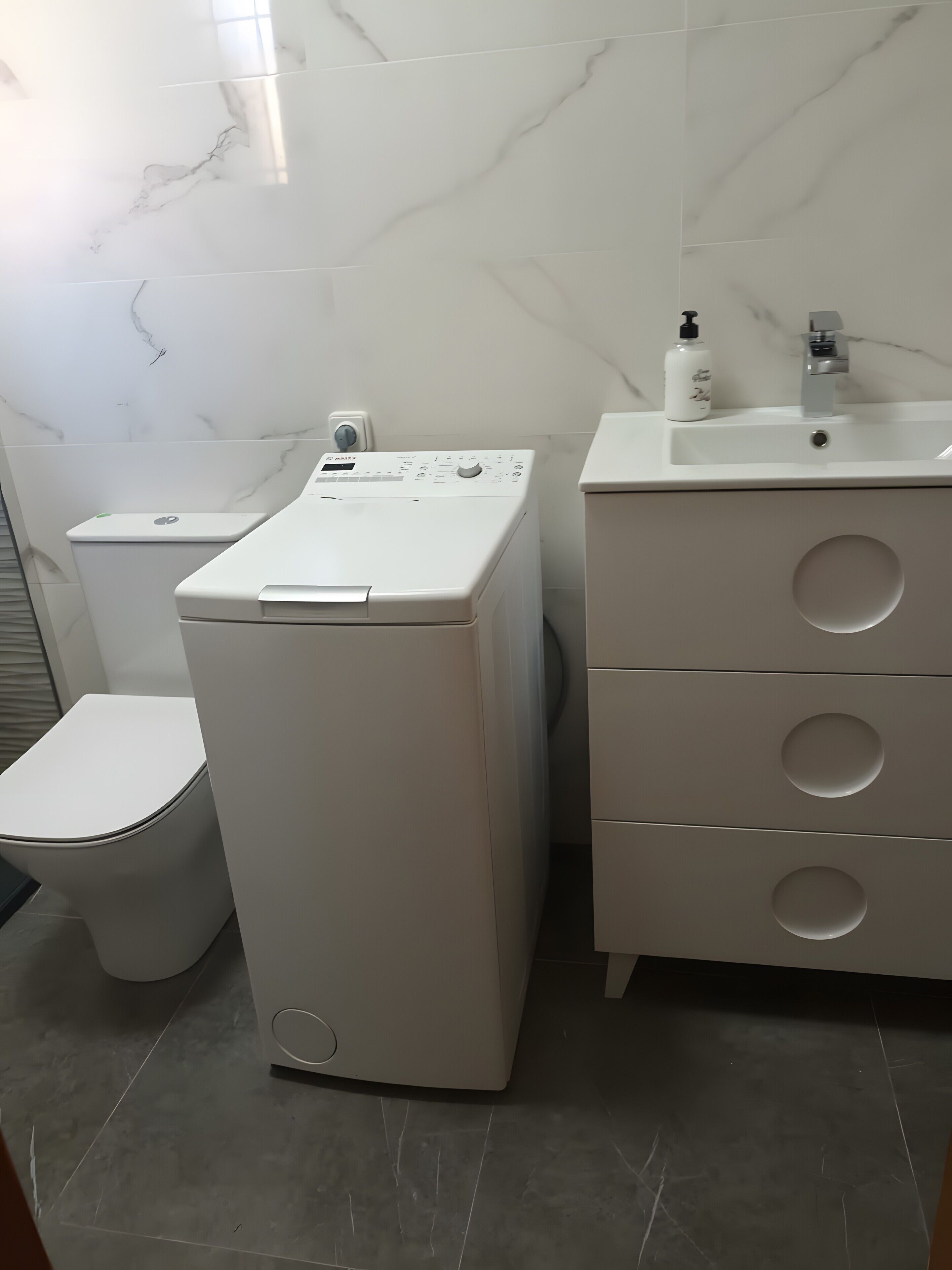 A compact laundry room with a front-loading washer, a toilet, and a sink with a mirror.