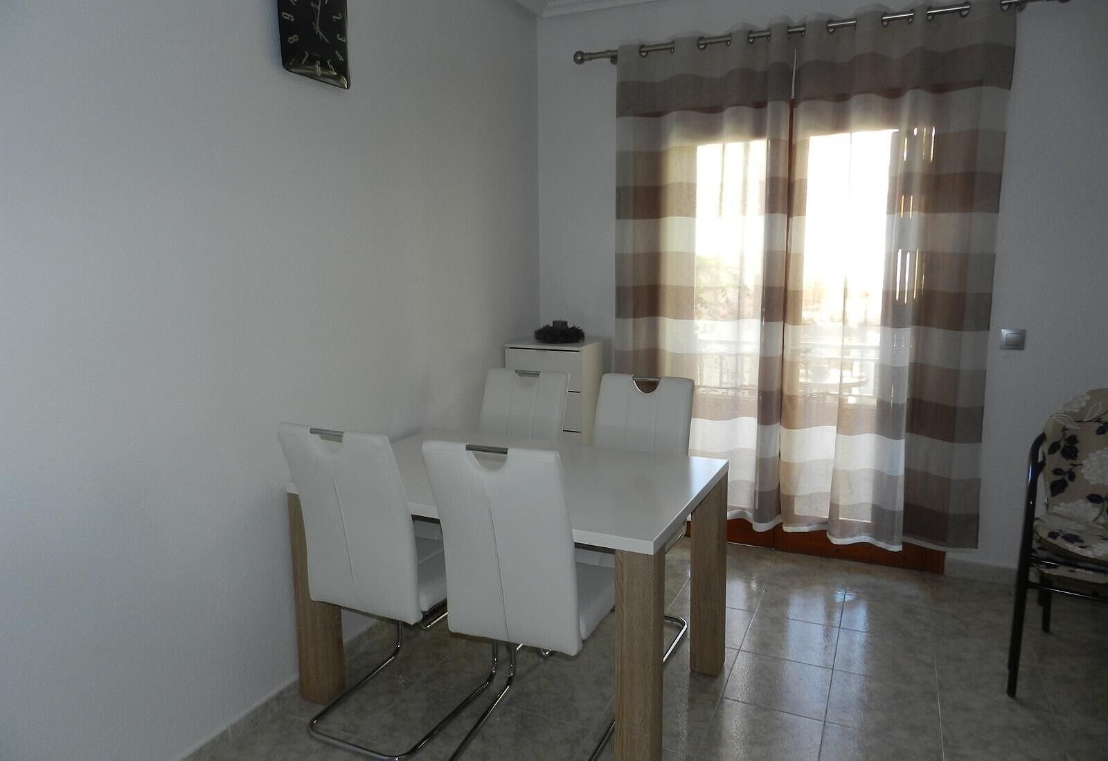 A dining area with a white table and chairs, a small cabinet, and a window with striped curtains.