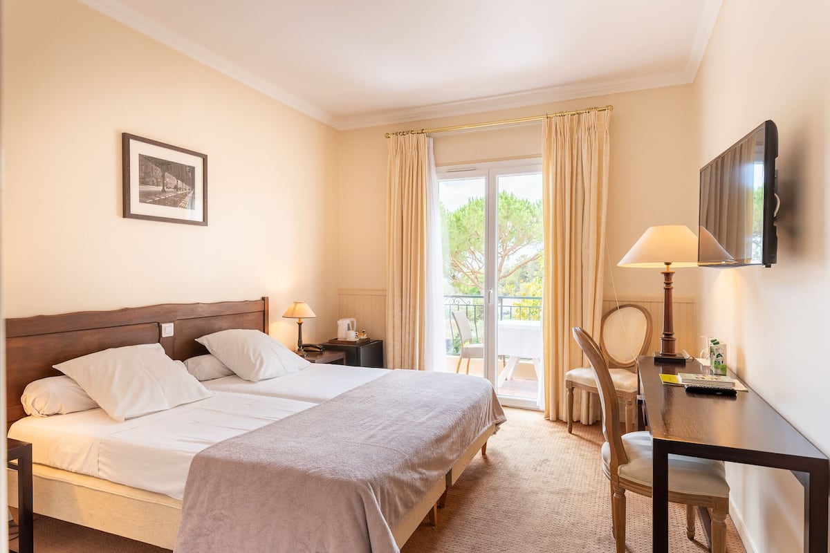 Habitación de hotel con cama, dos mesitas de noche, un escritorio con silla, televisor y balcón con cortinas.