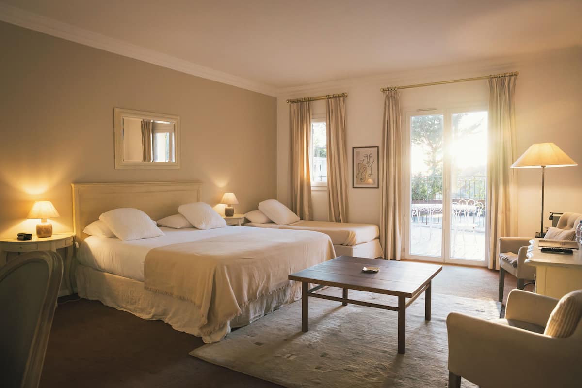 Habitación de hotel con una cama grande, una cama más pequeña, un escritorio y una silla. Se ve una ventana con cortinas y vistas al exterior.