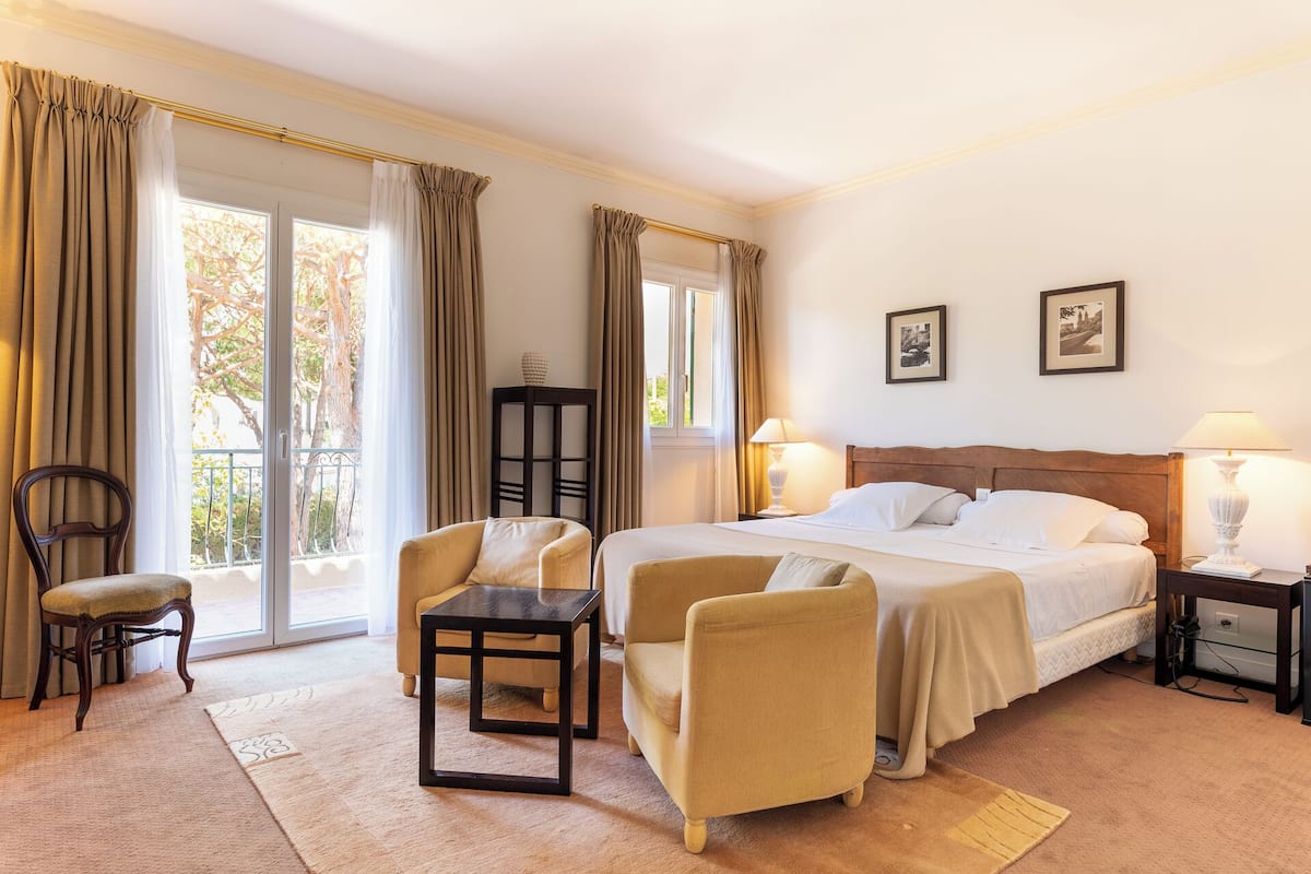 Habitación de hotel con cama, dos sillones, una mesita, una mesita de noche y ventana con cortinas.