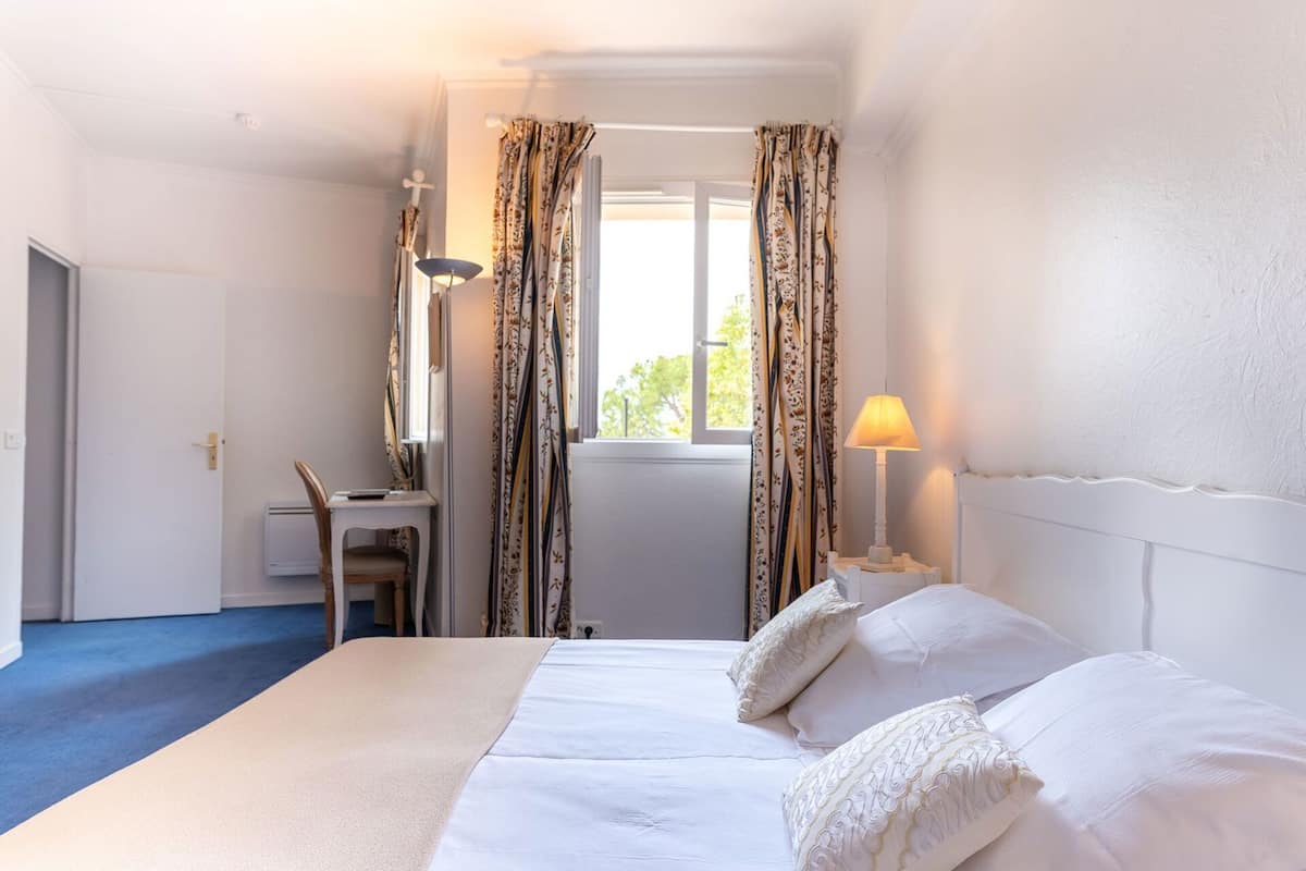 Habitación de hotel con cama, escritorio, lámpara y ventana con cortinas.