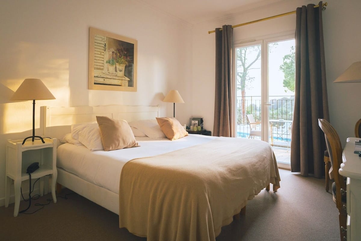Habitación de hotel con cama, dos lámparas de noche, mesita de noche, escritorio, silla, un cuadro en la pared y balcón con cortinas.