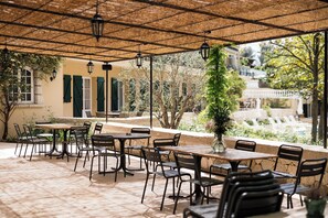 Un café al aire libre con mesas y sillas, una pérgola con faroles colgantes y vistas a una fuente y árboles.
