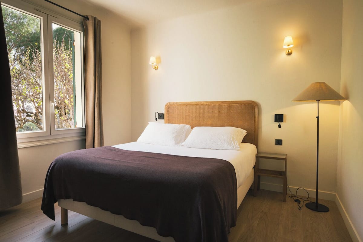 Un dormitorio con cama, lámparas de noche, una ventana con cortinas y vistas a árboles.