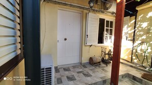 Un étroit couloir extérieur avec une porte blanche, une fenêtre avec des volets et une petite table surmontée d’un tonneau.