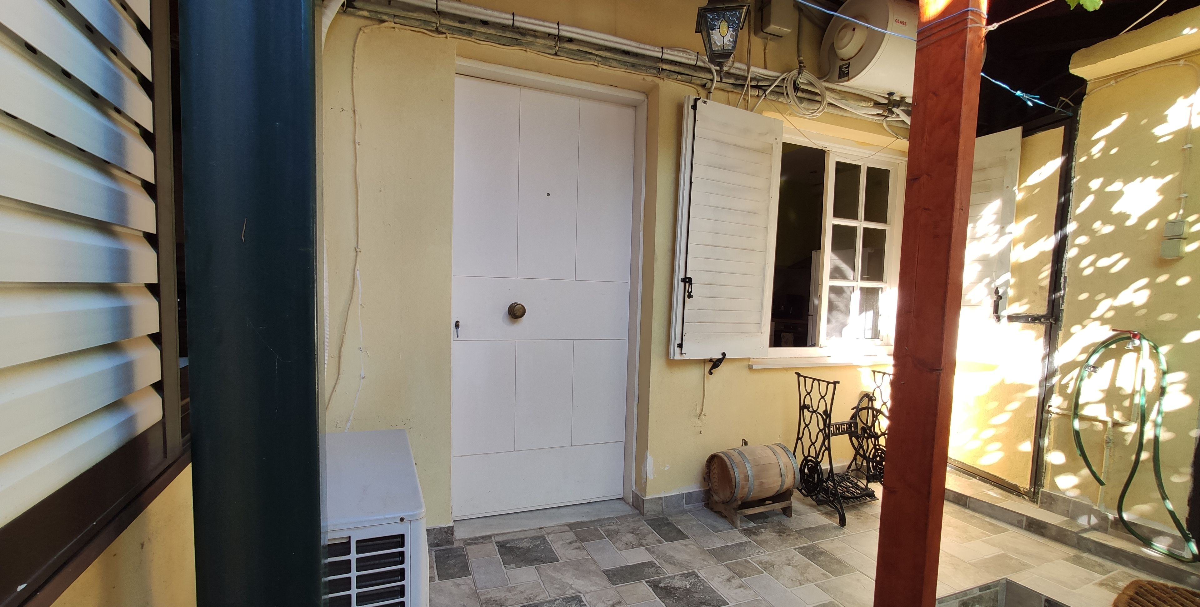 Un estrecho pasillo exterior con una puerta blanca, una ventana con contraventanas y una pequeña mesa con un barril.