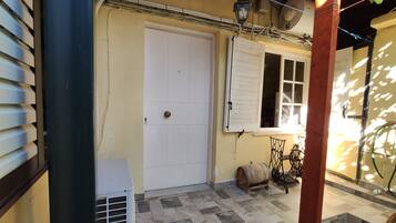 Un estrecho pasillo exterior con una puerta blanca, una ventana con contraventanas y una pequeña mesa con un barril.