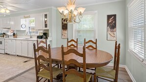 A dining area with a round wooden table and chairs, a chandelier, and a view of the kitchen in the background. - Byrd's Nest is located across the street from the beach and sleeps 10 (St Simons Island)