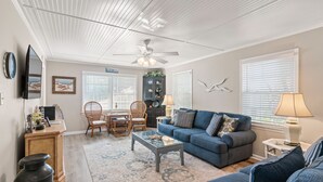 A living room with a blue sofa, a coffee table, a ceiling fan, and a dining area with chairs and a table. - Byrd's Nest is located across the street from the beach and sleeps 10 (St Simons Island)