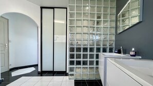 Een moderne badkamer met betegelde vloer, een glazen scheidingswand en een wit wastafelmeubel met zeepdispenser.