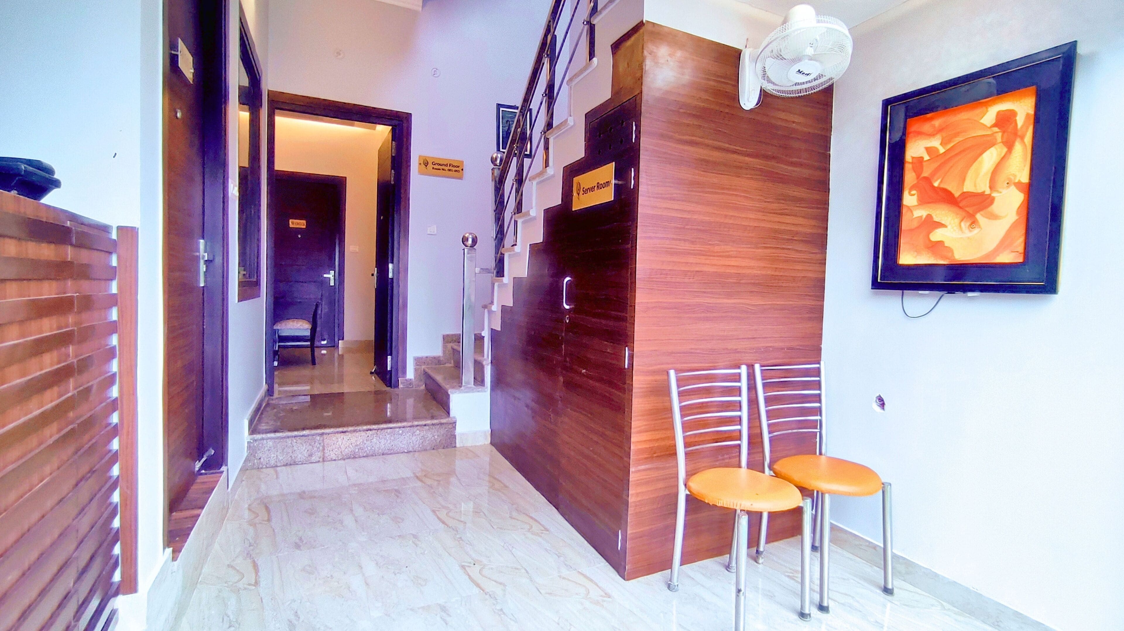 A hotel hallway with a wooden reception desk, two chairs, a server room sign, and a framed artwork on the wall.