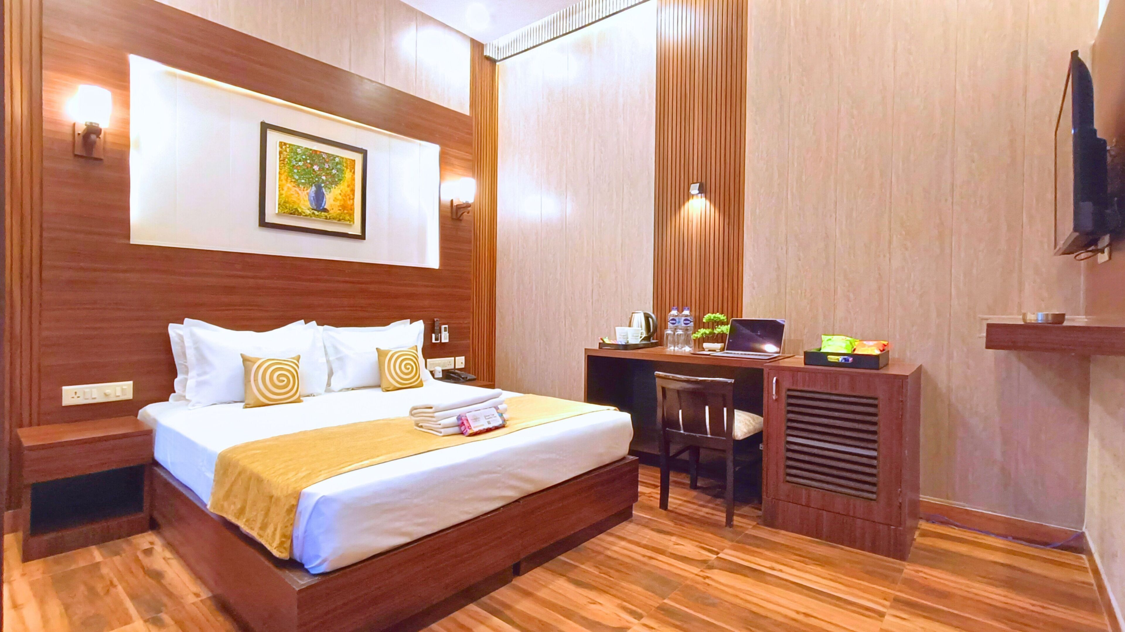 Habitación de hotel con una cama de madera, un escritorio con una computadora portátil, una silla y un televisor en la pared.