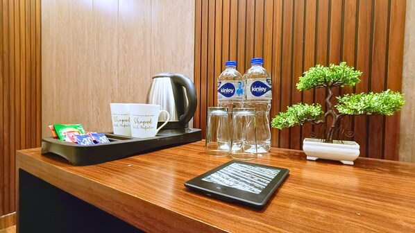 A wooden reception desk with a kettle, two water bottles, a tray with coffee cups, a small potted plant, and a tablet.