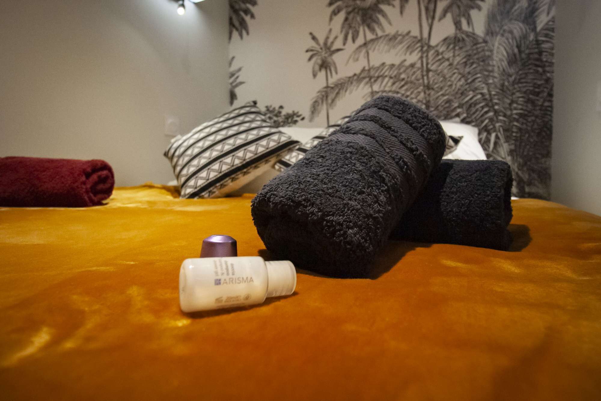 A bed with a yellow bedspread, a rolled-up black towel, a rolled-up dark towel, a white bottle labeled 'ARISMA', and a red towel.